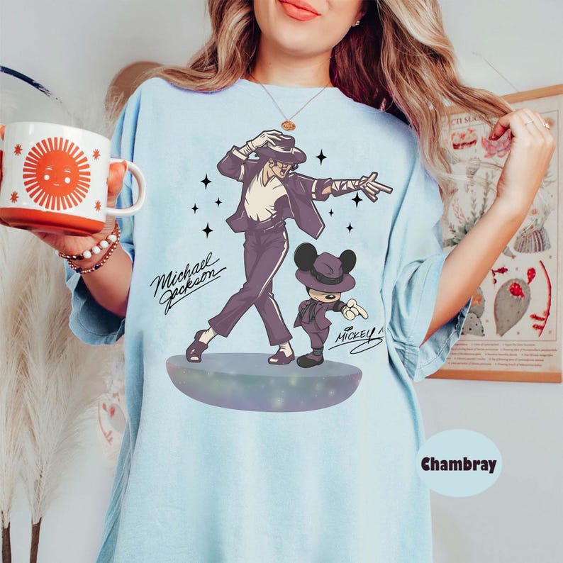 May include: Light blue t-shirt featuring a graphic of Michael Jackson and Mickey Mouse in matching outfits. The design includes the text "Michael Jackson" and "Mickey". The shirt is a light blue color.