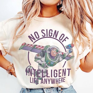 Pu&ograve; includere: T-shirt color crema con un'immagine a cartoni animati di Buzz Lightyear e la scritta "NO SIGN OF INTELLIGENT LIFE ANYWHERE". Buzz Lightyear &egrave; raffigurato nella sua tuta spaziale verde, bianca e viola, con una pistola a raggi blu.