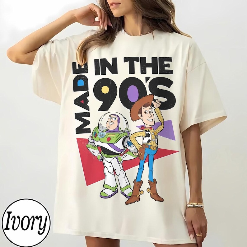May include: Ivory-colored t-shirt featuring a graphic of Buzz Lightyear and Woody from Toy Story, with the text "MADE IN THE 90'S". The design includes a red and purple geometric shape. The shirt has a relaxed fit.