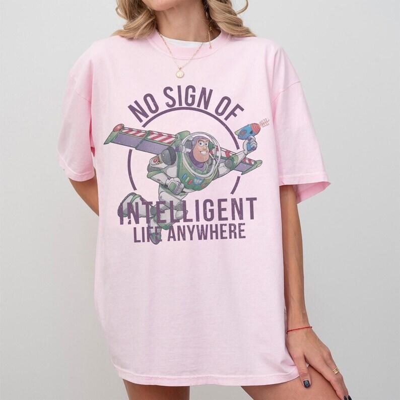 Pu&ograve; includere: T-shirt rosa chiaro con una grafica di Buzz Lightyear in volo, con una pistola laser. Il design include il testo "NO SIGN OF INTELLIGENT LIFE ANYWHERE" in un cerchio. La maglietta &egrave; di vestibilit&agrave; ampia.