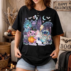 May include: A black short-sleeve t-shirt featuring a colorful graphic print of two cartoon characters, one pink and one blue, dressed for Halloween. The design includes a pumpkin, an ice cream cone, a castle, bats, and a crescent moon.