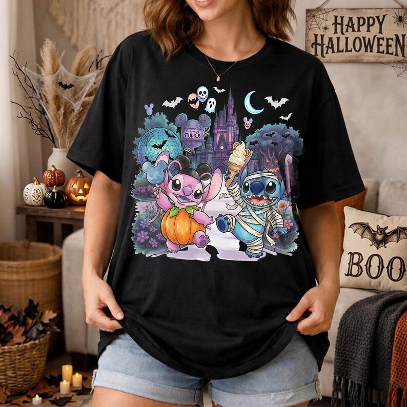May include: A black short-sleeve t-shirt featuring a colorful graphic print of two cartoon characters, one pink and one blue, dressed for Halloween. The design includes a pumpkin, an ice cream cone, a castle, bats, and a crescent moon.