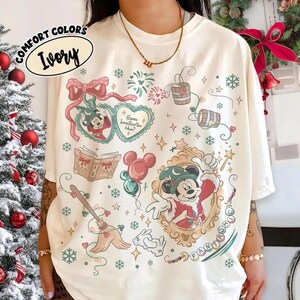 May include: An ivory t-shirt featuring a festive graphic print with Mickey Mouse in a wizard hat, a heart-shaped frame with the text Some Imagination Huh?, a book, a broom, balloons, and snowflakes. The shirt has a crew neckline and short sleeves.