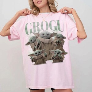 May include: A light pink t-shirt featuring the word "GROGU" in green above a graphic of Grogu from The Mandalorian. The graphic shows Grogu in various poses. The t-shirt has short sleeves and a relaxed fit.