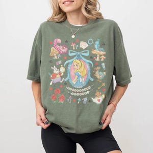 May include: Olive green t-shirt featuring a colorful Alice in Wonderland graphic. The design includes Alice, the White Rabbit, the Cheshire Cat, and other characters. The words "Wonderland" are also visible in the design.