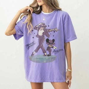 May include: A lilac-colored t-shirt featuring a graphic of Michael Jackson in a black suit and hat, alongside a Mickey Mouse character in a similar outfit. The text "Michael Jackson" and "Mickey Mouse" are also visible.