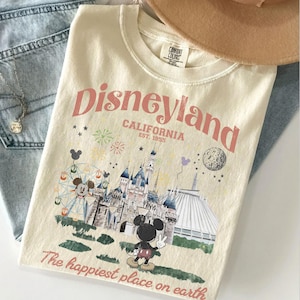 Puede incluir: Camiseta color crema con un gr&aacute;fico de Disneyland. El dise&ntilde;o incluye el logotipo de Disneyland, un castillo, Mickey Mouse y el texto "The happiest place on earth". La camiseta se combina con unos vaqueros azules y un sombrero marr&oacute;n.
