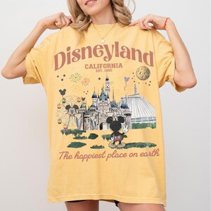 Puede incluir: Camiseta amarilla con un dise&ntilde;o de Disneyland. El dise&ntilde;o incluye el logotipo de Disneyland, la ubicaci&oacute;n en California y el a&ntilde;o de establecimiento. El dise&ntilde;o tambi&eacute;n muestra un castillo, Mickey Mouse y la frase "The happiest place on earth."