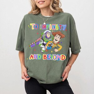 May include: An olive green t-shirt featuring the phrase "TO INFINITY AND BEYOND" in colorful text. The shirt displays a graphic of Buzz Lightyear and Woody from Toy Story. The shirt is a short-sleeved, crew neck style.