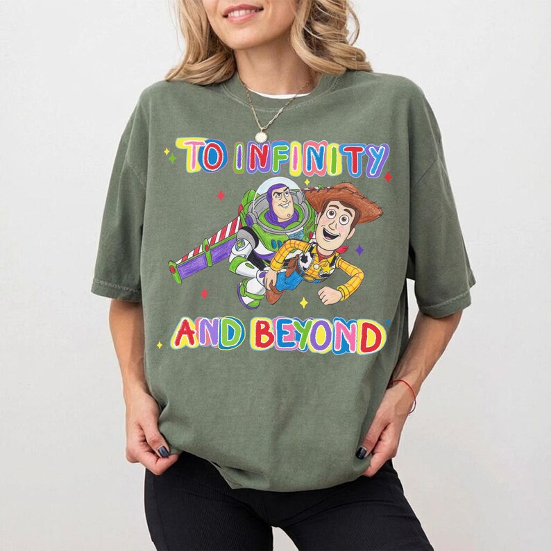 May include: An olive green t-shirt featuring the phrase "TO INFINITY AND BEYOND" in colorful text. The shirt displays a graphic of Buzz Lightyear and Woody from Toy Story. The shirt is a short-sleeved, crew neck style.