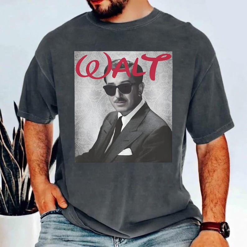May include: Dark gray t-shirt featuring a black and white image of a man wearing sunglasses and a suit. The word "WALT" is printed in red cursive above the image. The shirt is made of a soft material.
