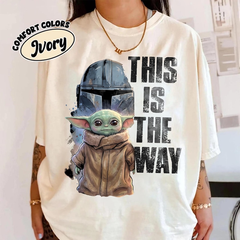 May include: Ivory t-shirt with a graphic of a Mandalorian helmet and Grogu, and the text "THIS IS THE WAY." The shirt also has the text "COMFORT COLORS" and "Ivory" in a circular design.