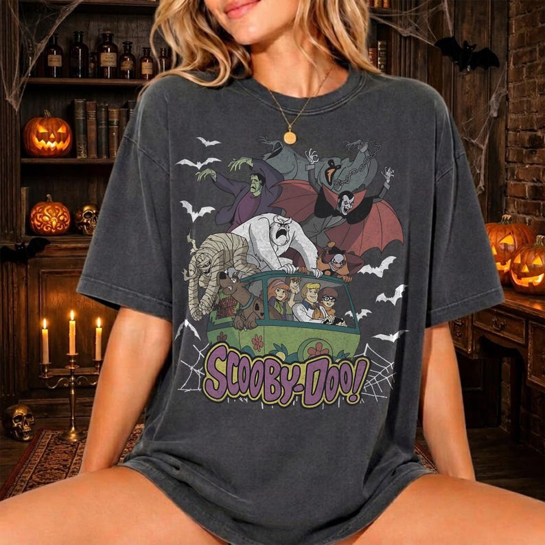 May include: A charcoal gray short-sleeve t-shirt featuring a graphic print of the Scooby-Doo characters in a green van surrounded by monsters, bats, and spiderwebs. The text "SCOOBY-DOO!" is printed in purple and yellow below the illustration.