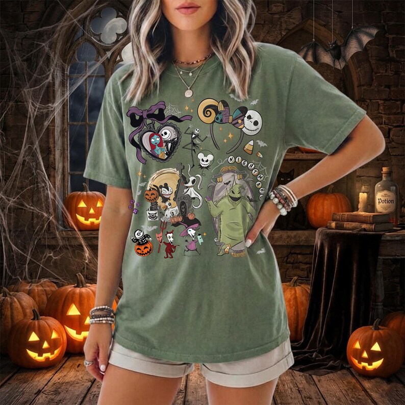 May include: A sage green short-sleeve t-shirt featuring a graphic print of characters from The Nightmare Before Christmas, including Jack Skellington, Sally, and Oogie Boogie, alongside festive icons like pumpkins and mouse-ear headbands.