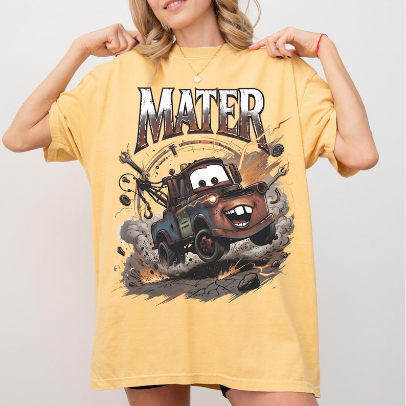 May include: Yellow t-shirt featuring the word "MATER" in a distressed font above a cartoon image of Mater the tow truck from the movie Cars. The graphic shows Mater kicking up dust and debris.