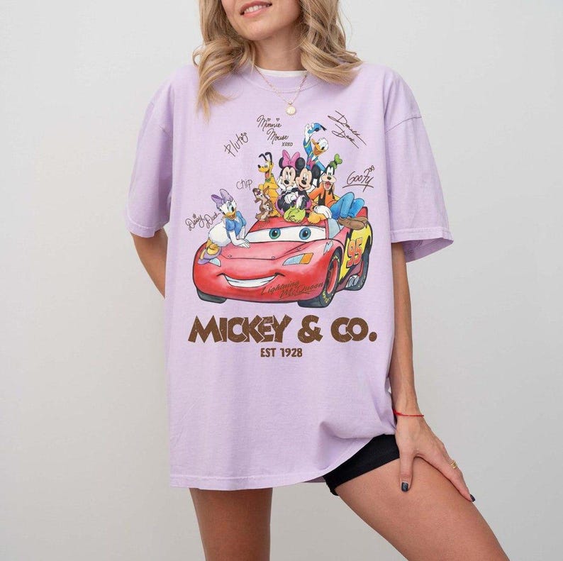 May include: A light purple t-shirt featuring a graphic of Disney characters in a red car. The text "Mickey & Co. Est 1928" is printed below the graphic. Signatures of the characters are above the car.