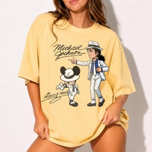K&ouml;nnte beinhalten: Gelbes kurz&auml;rmeliges T-Shirt mit einer Cartoon-Illustration von Michael Jackson und Mickey Mouse in passenden Outfits. Das Shirt hat den Text "Michael Jackson" und "Mickey Mouse" in einer Schriftart.