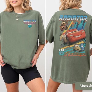 May include: An olive green t-shirt featuring a small Radiator Springs US 66 logo on the front chest. The back displays a large graphic of Lightning McQueen, Mater, and other characters with the text Radiator Springs and a US 66 road sign.