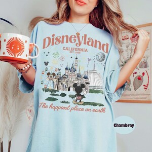 Puede incluir: Camiseta azul claro con un gr&aacute;fico de Disneyland. El dise&ntilde;o presenta el logotipo de Disneyland, un castillo, Mickey Mouse y el texto "The happiest place on earth". Se sostiene una taza blanca y naranja con un dise&ntilde;o de sol.