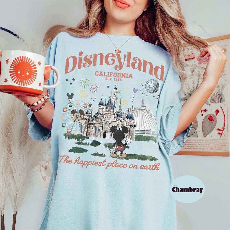 Puede incluir: Camiseta azul claro con un gr&aacute;fico de Disneyland. El dise&ntilde;o presenta el logotipo de Disneyland, un castillo, Mickey Mouse y el texto "The happiest place on earth". Se sostiene una taza blanca y naranja con un dise&ntilde;o de sol.