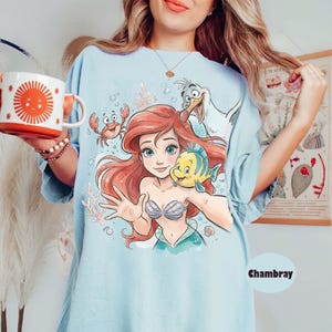 Op de afbeelding: Een lichtblauw t-shirt met een grafische print van een zeemeermin met rood haar, een geel en blauwe vis, een rode krab en een witte meeuw. Het ontwerp bevat koraal en bubbels. Het t-shirt is gelabeld als chambray en heeft een ronde hals.