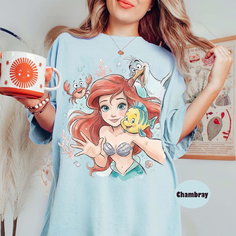 Op de afbeelding: Een lichtblauw t-shirt met een grafische print van een zeemeermin met rood haar, een geel en blauwe vis, een rode krab en een witte meeuw. Het ontwerp bevat koraal en bubbels. Het t-shirt is gelabeld als chambray en heeft een ronde hals.