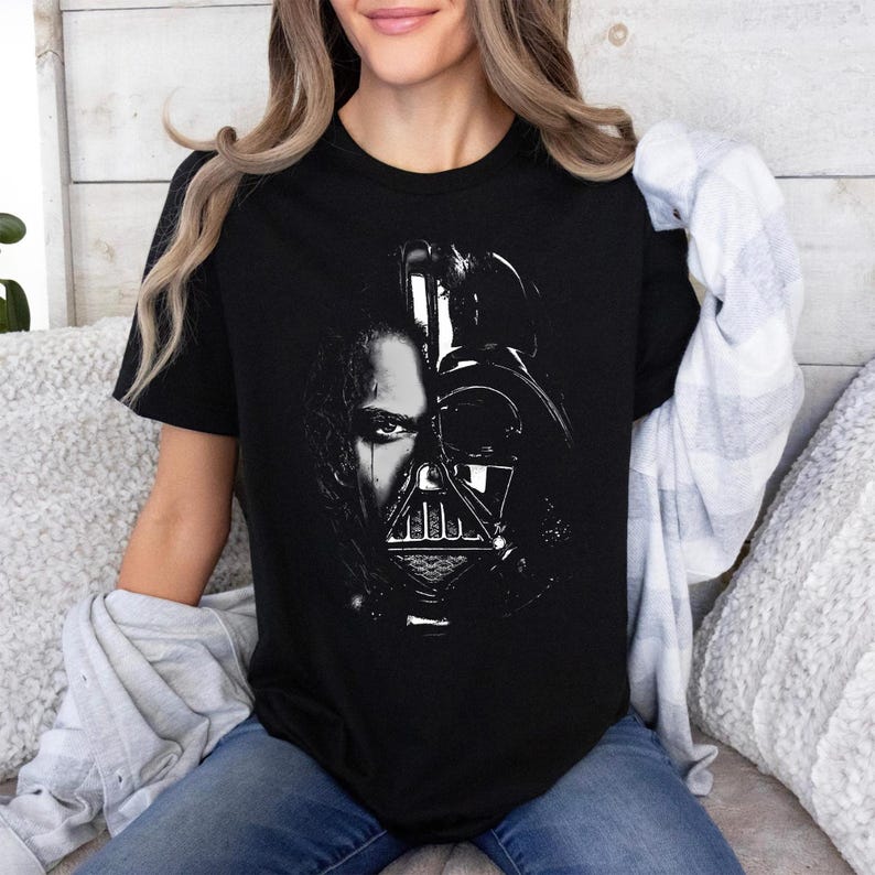 May include: Black t-shirt featuring a split image of Darth Vader and a human face. The graphic is in black and white, with the human face on the left and Vader's helmet on the right. The shirt is short-sleeved and worn by a person.