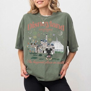 Puede incluir: Camiseta verde oliva con un gr&aacute;fico de Disneyland. El gr&aacute;fico presenta un castillo, Space Mountain, una noria y Mickey Mouse. El texto de la camiseta dice "Disneyland California Est. 1955" y "The happiest place on earth."