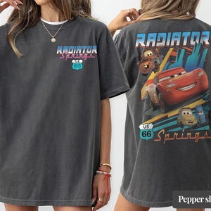 May include: A charcoal gray short-sleeve t-shirt featuring a small Radiator Springs U.S. 66 logo on the front left chest. The back displays a large graphic of Lightning McQueen, Mater, and other characters with the text Radiator Springs U.S. 66.