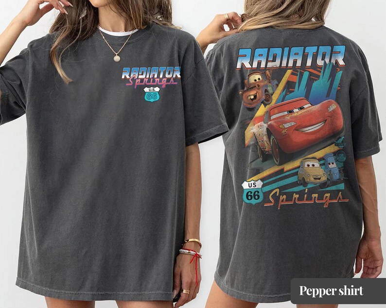 May include: A charcoal gray short-sleeve t-shirt featuring a small Radiator Springs U.S. 66 logo on the front left chest. The back displays a large graphic of Lightning McQueen, Mater, and other characters with the text Radiator Springs U.S. 66.