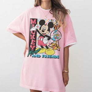 May include: A light pink oversized t-shirt featuring Mickey Mouse and friends. The graphic includes Mickey Mouse, Donald Duck, and Pluto, with the text "Mickey Mouse and Friends". The shirt has white trim on the sleeves.