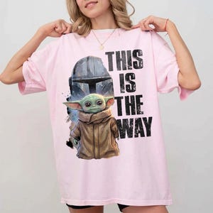 May include: A pale pink t-shirt with a graphic of a Mandalorian helmet and Grogu, with the text "THIS IS THE WAY" in black. The design includes a detailed illustration of Grogu in a brown coat. The shirt has a relaxed, oversized fit.