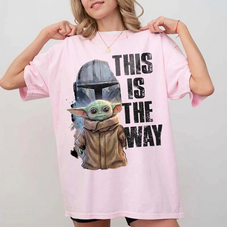 May include: A pale pink t-shirt with a graphic of a Mandalorian helmet and Grogu, with the text "THIS IS THE WAY" in black. The design includes a detailed illustration of Grogu in a brown coat. The shirt has a relaxed, oversized fit.