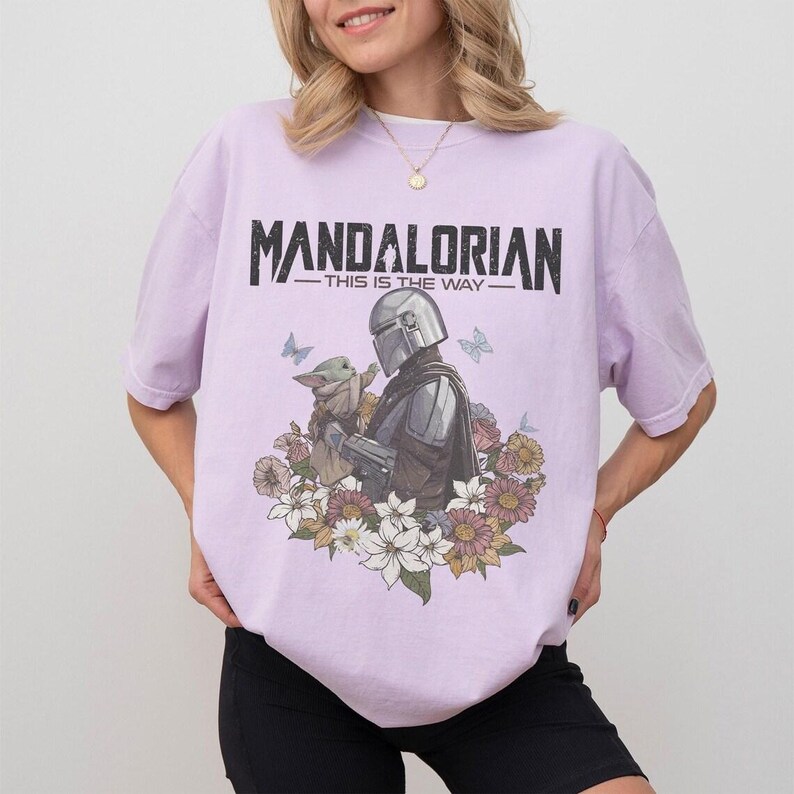 K&ouml;nnte beinhalten: Helllila T-Shirt mit einem Aufdruck von The Mandalorian und Grogu, umgeben von Blumen und Schmetterlingen. Der Text "MANDALORIAN - THIS IS THE WAY" ist schwarz gedruckt. Das T-Shirt hat eine lockere Passform.