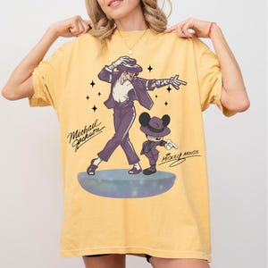 May include: Mustard yellow t-shirt featuring a graphic of Michael Jackson and Mickey Mouse in matching purple outfits. The text "Michael Jackson" and "Mickey Mouse" are written in cursive. The design includes stars and a stage.