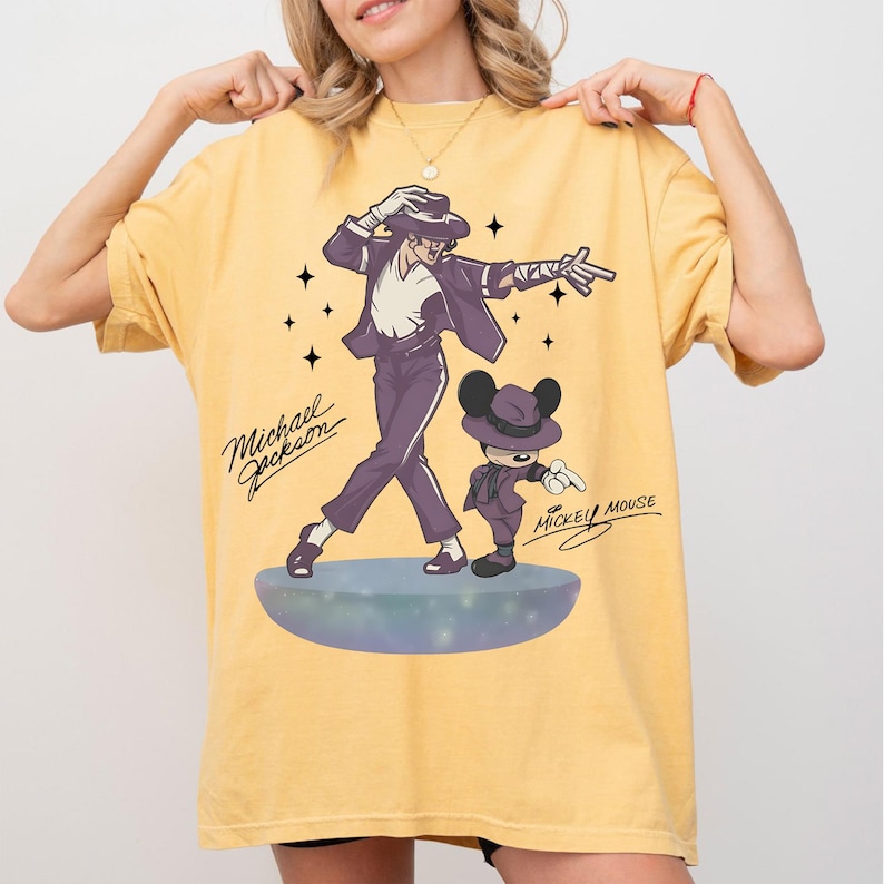 May include: Mustard yellow t-shirt featuring a graphic of Michael Jackson and Mickey Mouse in matching purple outfits. The text "Michael Jackson" and "Mickey Mouse" are written in cursive. The design includes stars and a stage.