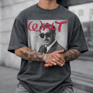 May include: Dark gray t-shirt featuring a black and white image of a man wearing sunglasses, with the name "WALT" in red script above the image. The shirt has short sleeves and a relaxed fit.