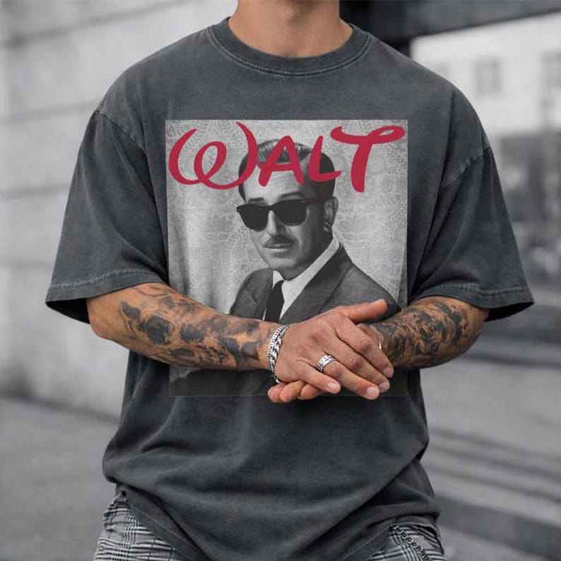 May include: Dark gray t-shirt featuring a black and white image of a man wearing sunglasses, with the name "WALT" in red script above the image. The shirt has short sleeves and a relaxed fit.