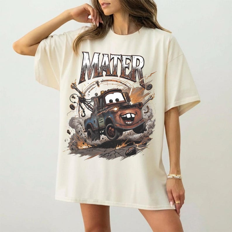 May include: Cream-colored t-shirt featuring a graphic of Mater from the movie Cars. The design shows Mater in action, with the word "MATER" above. The shirt has short sleeves and a relaxed fit.