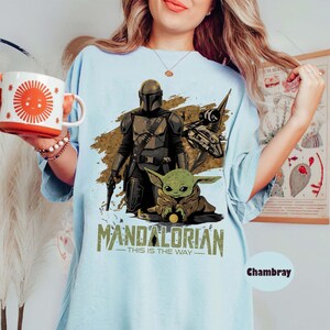 May include: Light blue t-shirt featuring a graphic of The Mandalorian, Grogu, and a spaceship. The text "MANDALORIAN - THIS IS THE WAY" is printed below the characters. The shirt is a light blue color.