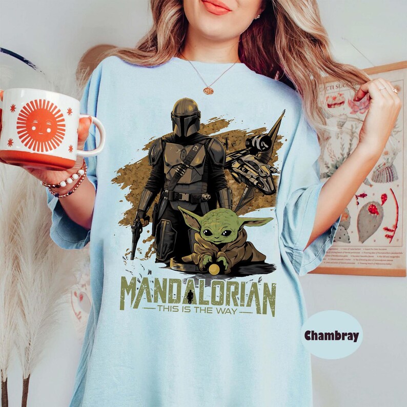 May include: Light blue t-shirt featuring a graphic of The Mandalorian, Grogu, and a spaceship. The text "MANDALORIAN - THIS IS THE WAY" is printed below the characters. The shirt is a light blue color.