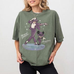 May include: Olive green t-shirt featuring a graphic of Michael Jackson and Mickey Mouse in matching outfits. The design includes the text "Michael Jackson" and "Mickey Mouse" in a stylized font. The shirt has short sleeves.