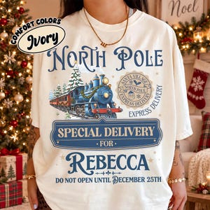 May include: An ivory t-shirt featuring a graphic of a blue steam train, a circular North Pole Santa Village Express Delivery stamp, and text that reads North Pole, Special Delivery for Rebecca, and Do Not Open Until December 25th.