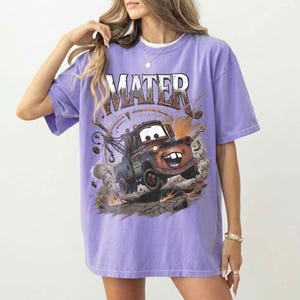 May include: A purple t-shirt featuring the word "MATER" in a distressed font above a cartoon image of Mater from the movie Cars. The image shows Mater as a tow truck kicking up dust and debris. The shirt is a relaxed fit.