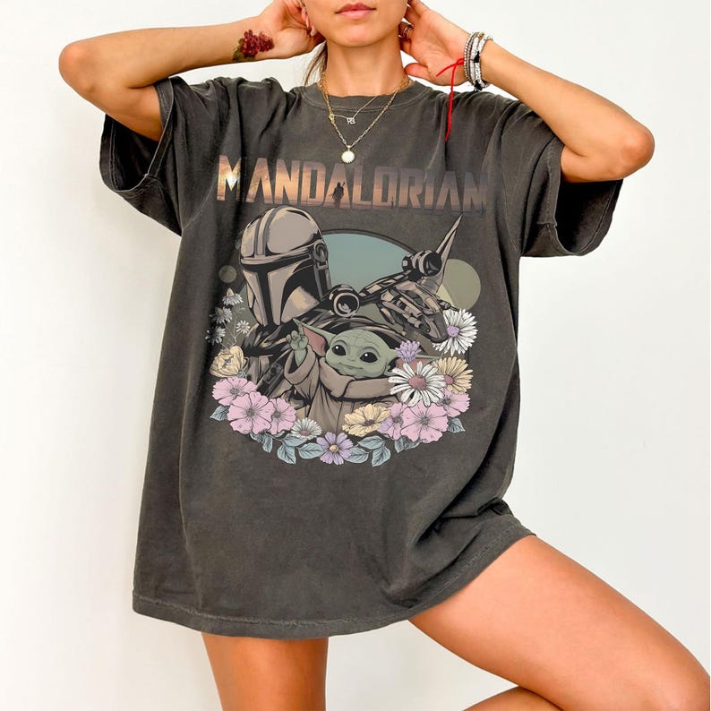 May include: Dark gray t-shirt featuring the word "MANDALORIAN" above a graphic of the Mandalorian, Grogu, and a ship, surrounded by flowers. The shirt has short sleeves and a relaxed fit.
