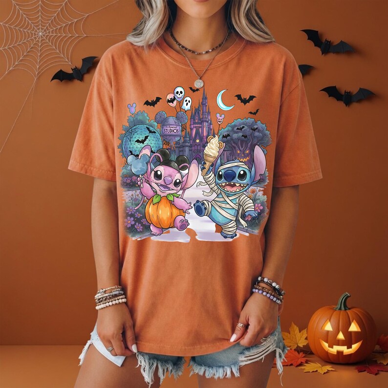 May include: An orange short-sleeve t-shirt featuring a graphic print of two blue and pink alien characters in Halloween costumes. One character is dressed as a pumpkin and the other as a mummy, standing before a castle with bats and balloons.