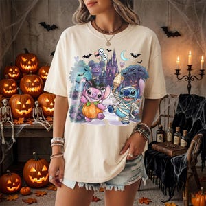 May include: A cream-colored t-shirt featuring a graphic print of two cartoon characters, one pink and one blue, dressed in Halloween costumes. The design includes a pumpkin, a castle, bats, and a crescent moon. The characters are holding balloons.
