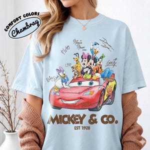 May include: Light blue t-shirt featuring a red Lightning McQueen car with Disney characters. The shirt includes the text "Mickey & Co. EST 1928" and signatures of the characters. The shirt is made of Comfort Colors Chambray material.