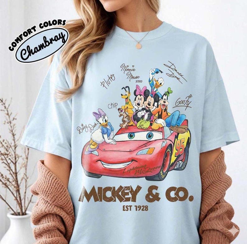 May include: Light blue t-shirt featuring a red Lightning McQueen car with Disney characters. The shirt includes the text "Mickey & Co. EST 1928" and signatures of the characters. The shirt is made of Comfort Colors Chambray material.