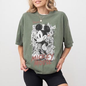 Puede incluir: Camiseta verde oliva con un gr&aacute;fico en blanco y negro de Mickey Mouse. El dise&ntilde;o incluye las palabras "Mickey Mouse" en rojo y un fondo de paisaje urbano. La camiseta tiene mangas cortas y un corte holgado.